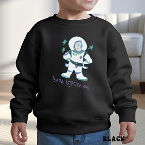 May include: Black sweatshirt featuring a cartoon illustration of Buzz Lightyear in a white spacesuit with green and purple accents. The text "Buzz Lightyear" is written in purple script below the character. The word "BLACK" is printed in white at the bottom.