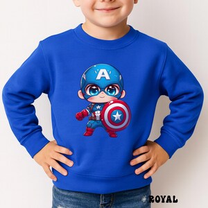Op de afbeelding: Koningsblauw sweatshirt met een cartoon Captain America-afbeelding. De afbeelding toont Captain America met een blauwe helm met een witte "A", die een schild met een ster vasthoudt. De sweatshirt heeft een ronde hals.