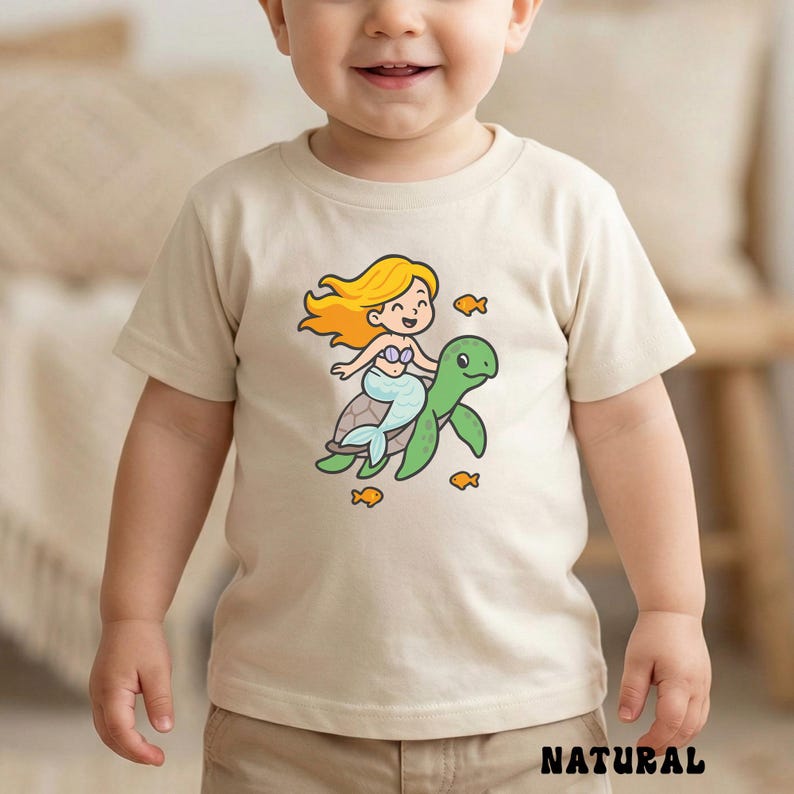 Puede incluir: Camiseta beige con una sirena de dibujos animados montada en una tortuga verde, rodeada de peque&ntilde;os peces naranjas. La sirena tiene pelo rubio y una cola azul. La palabra "NATURAL" est&aacute; impresa en la parte inferior de la camiseta.