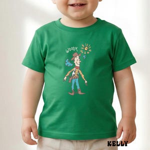 May include: A green t-shirt featuring a cartoon illustration of Woody from Toy Story. The design includes Woody in his cowboy attire, the word "WOODY", and a sun graphic. The shirt has short sleeves and is worn by a child.