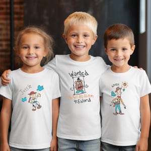 May include: Three children wearing white t-shirts with cartoon character designs. One shirt says "Jessie" with a drawing of a cowgirl. Another says "Wanted" with a Mr. Potato Head drawing. The third says "Woody" with a cowboy drawing.