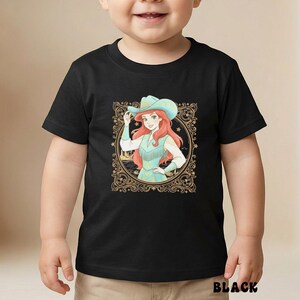 K&ouml;nnte beinhalten: Schwarzes T-Shirt mit einer Cartoon-Illustration einer Cowgirl mit roten Haaren, die einen t&uuml;rkisfarbenen Hut und eine Weste tr&auml;gt. Das Design ist in einem goldenen, verzierten Rahmen eingefasst. Das Wort "BLACK" ist unten aufgedruckt.