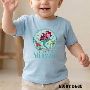 May include: Light blue t-shirt featuring a graphic from Disney's The Little Mermaid. The design includes Ariel, Flounder, and Sebastian, surrounded by aquatic plants. The shirt has the text "The Little Mermaid" below the graphic.