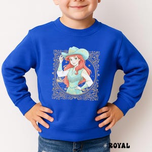 K&ouml;nnte beinhalten: K&ouml;nigsblaues Sweatshirt mit einer Cartoon-Illustration einer Figur mit Cowboyhut und Fransenweste. Die Grafik ist von einem goldenen Zierrahmen mit Sternen umgeben. Das Wort "ROYAL" ist unten aufgedruckt.
