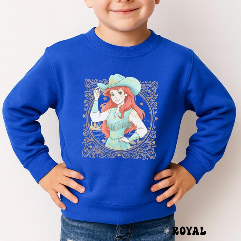 K&ouml;nnte beinhalten: K&ouml;nigsblaues Sweatshirt mit einer Cartoon-Illustration einer Figur mit Cowboyhut und Fransenweste. Die Grafik ist von einem goldenen Zierrahmen mit Sternen umgeben. Das Wort "ROYAL" ist unten aufgedruckt.