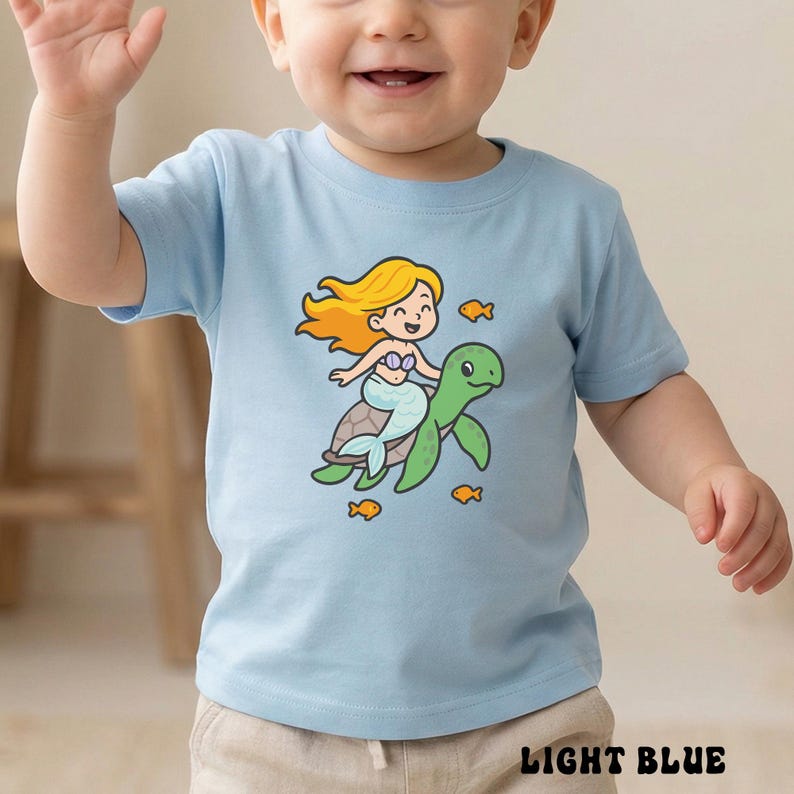 Puede incluir: Camiseta azul claro para ni&ntilde;os peque&ntilde;os con una sirena de dibujos animados montada en una tortuga verde, rodeada de peque&ntilde;os peces naranjas. La sirena tiene pelo largo y amarillo y una cara sonriente. El texto "LIGHT BLUE" est&aacute; impreso en la parte inferior de la camiseta.