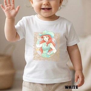 K&ouml;nnte beinhalten: Wei&szlig;es kurz&auml;rmeliges T-Shirt mit einer Cartoon-Illustration einer Figur mit Cowboyhut und Kleid. Die Illustration ist in einem goldfarbenen, verzierten Rahmen eingefasst. Das Wort "WHITE" ist unten in Schwarz gedruckt.
