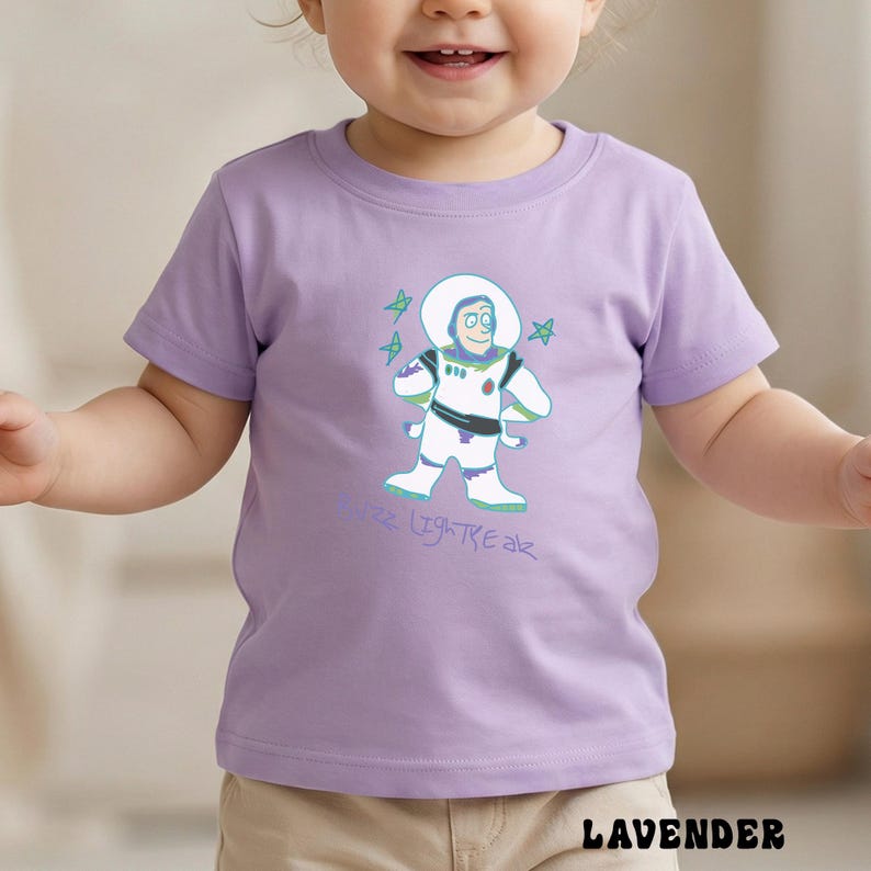 May include: A lavender short-sleeved t-shirt featuring a cartoon illustration of Buzz Lightyear in his space suit. The design includes the text "Buzz Lightyear" in a light blue color, along with green stars. The shirt is made of a soft material.