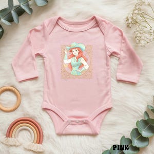 K&ouml;nnte beinhalten: Ein rosa Baby-Body mit langen &Auml;rmeln, der eine Cartoon-Cowgirl-Illustration zeigt. Die Illustration zeigt eine Frau mit roten Haaren, die einen t&uuml;rkisfarbenen Hut und ein Kleid tr&auml;gt. Der Body ist mit dekorativen Elementen verziert.