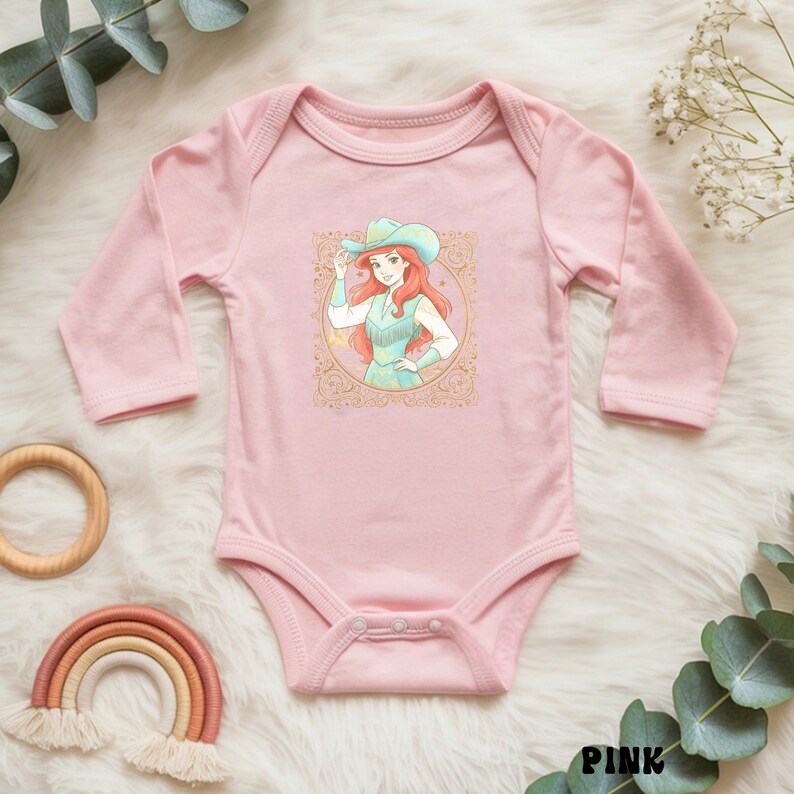K&ouml;nnte beinhalten: Ein rosa Baby-Body mit langen &Auml;rmeln, der eine Cartoon-Cowgirl-Illustration zeigt. Die Illustration zeigt eine Frau mit roten Haaren, die einen t&uuml;rkisfarbenen Hut und ein Kleid tr&auml;gt. Der Body ist mit dekorativen Elementen verziert.