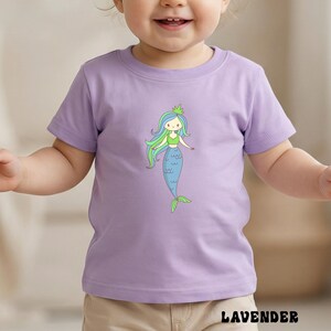 Puede incluir: Camiseta de color lavanda con una ilustraci&oacute;n de sirena de dibujos animados. La sirena tiene pelo azul y verde, un top verde y una cola azul. La palabra "LAVENDER" est&aacute; impresa en la parte inferior de la camiseta.