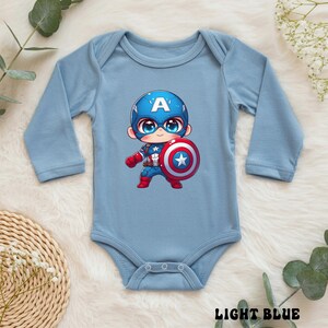 Op de afbeelding: Lichtblauwe babybody met een Captain America cartoonontwerp. De illustratie toont Captain America met zijn schild. De body heeft lange mouwen en de woorden "LIGHT BLUE" staan onderaan gedrukt.