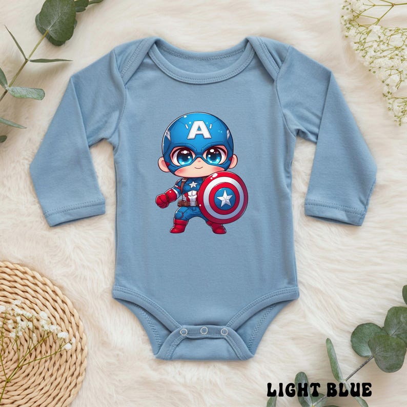 Op de afbeelding: Lichtblauwe babybody met een Captain America cartoonontwerp. De illustratie toont Captain America met zijn schild. De body heeft lange mouwen en de woorden "LIGHT BLUE" staan onderaan gedrukt.