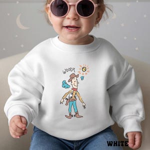 May include: White sweatshirt featuring a cartoon illustration of Woody from Toy Story. The design includes the word "WOODY" above the character, with a sun and cloud graphic. The word "WHITE" is printed at the bottom.
