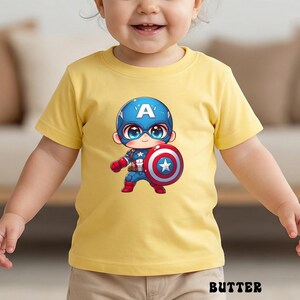 Op de afbeelding: Een gele t-shirt met een cartoon Captain America-afbeelding. De illustratie toont Captain America in een blauwe helm met een witte "A", die zijn schild vasthoudt. Het woord "BUTTER" staat onderaan het shirt.