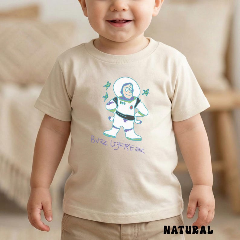 May include: A natural-colored t-shirt featuring a cartoon illustration of Buzz Lightyear from Toy Story. The design includes Buzz in his spacesuit, with the text "Buzz Lightyear" in a playful font. The word "NATURAL" is printed at the bottom.