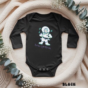 May include: Black baby bodysuit with long sleeves. The bodysuit features a cartoon graphic of Buzz Lightyear in a white spacesuit with green and purple accents. The text "Buzz Lightyear" is written in purple script below the graphic. The word "BLACK" is at the bottom.