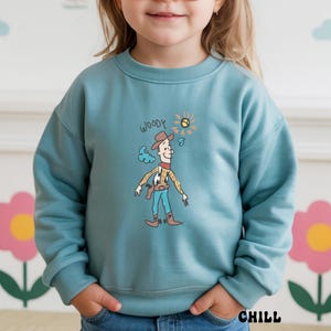 May include: A teal sweatshirt featuring a cartoon illustration of Woody from Toy Story. The design includes Woody in his cowboy attire, a sun, and the word "WOODY" above the character. The word "CHILL" is printed at the bottom.
