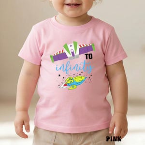 May include: Pink short-sleeved t-shirt with a colorful graphic design. The design includes a space ship, the words "TO infinity" in blue script, and a space ship. The word "PINK" is printed at the bottom.