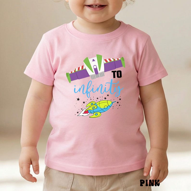 May include: Pink short-sleeved t-shirt with a colorful graphic design. The design includes a space ship, the words "TO infinity" in blue script, and a space ship. The word "PINK" is printed at the bottom.