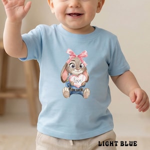Peut inclure: T-shirt bleu clair pour tout-petits avec une illustration &agrave; l'aquarelle d'un lapin tenant un c&oelig;ur avec le mot "Love". Le lapin porte un n&oelig;ud rose et un jean bleu. Les mots "LIGHT BLUE" sont imprim&eacute;s en bas du t-shirt.