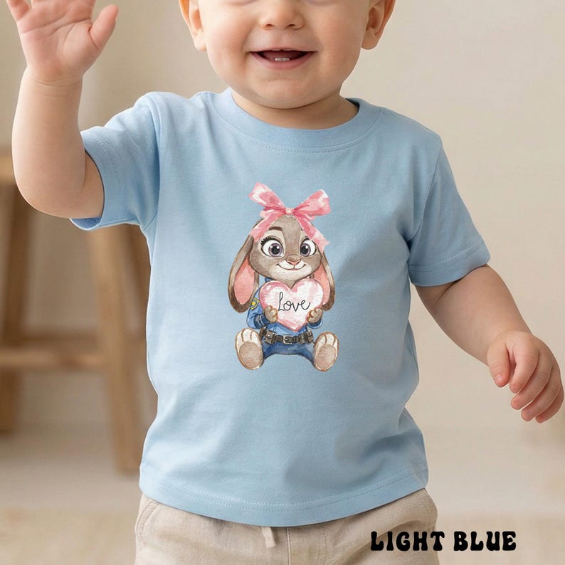 Peut inclure: T-shirt bleu clair pour tout-petits avec une illustration &agrave; l'aquarelle d'un lapin tenant un c&oelig;ur avec le mot "Love". Le lapin porte un n&oelig;ud rose et un jean bleu. Les mots "LIGHT BLUE" sont imprim&eacute;s en bas du t-shirt.