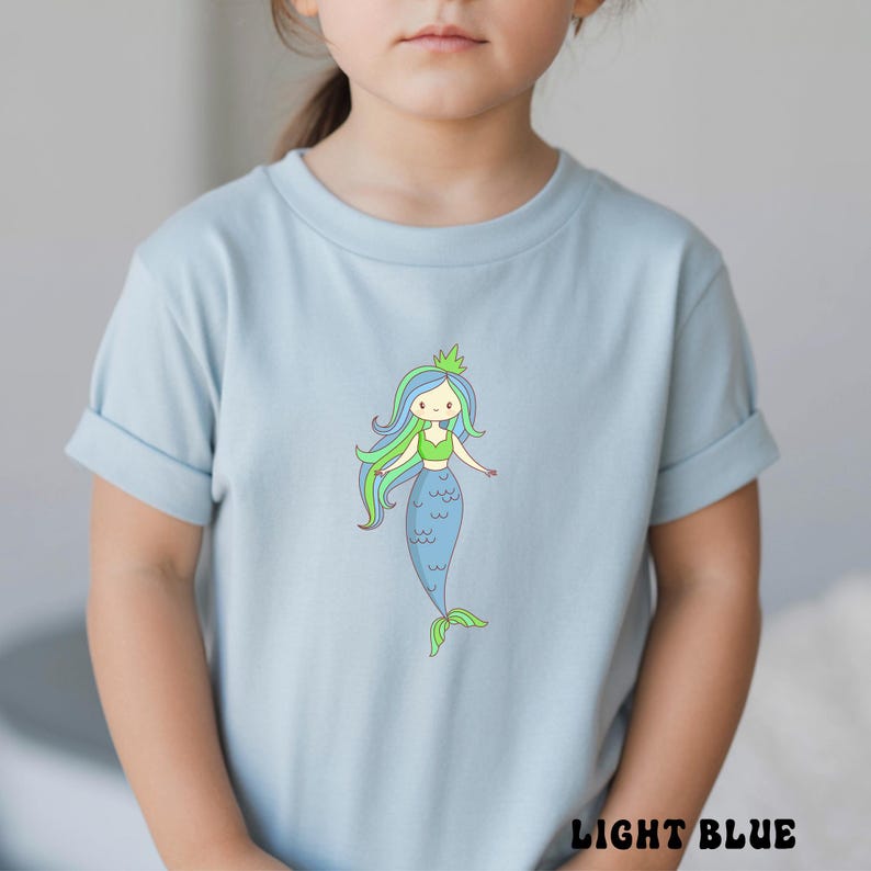 Puede incluir: Camiseta azul claro con una ilustraci&oacute;n de sirena de dibujos animados. La sirena tiene cabello azul y verde, un top verde y una cola azul. El texto "LIGHT BLUE" est&aacute; en la parte inferior de la camiseta.