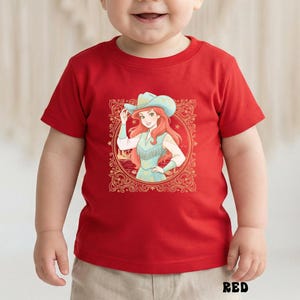K&ouml;nnte beinhalten: Rotes Kleinkind-T-Shirt mit einer Grafik einer Cowgirl mit roten Haaren und einem blauen Hut. Das Cowgirl befindet sich in einem dekorativen Rahmen. Das Wort "RED" ist unten auf dem Shirt in Schwarz gedruckt.