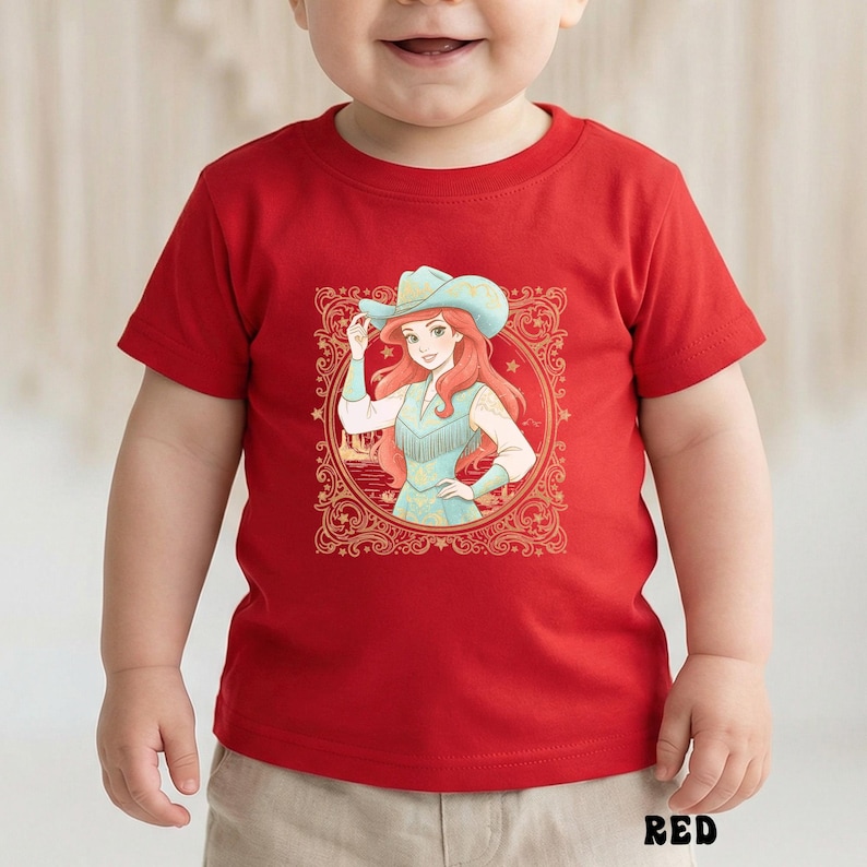 K&ouml;nnte beinhalten: Rotes Kleinkind-T-Shirt mit einer Grafik einer Cowgirl mit roten Haaren und einem blauen Hut. Das Cowgirl befindet sich in einem dekorativen Rahmen. Das Wort "RED" ist unten auf dem Shirt in Schwarz gedruckt.