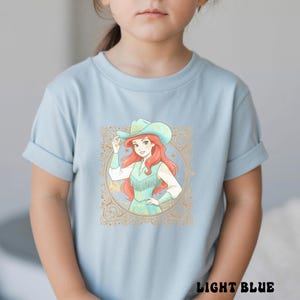 K&ouml;nnte beinhalten: Hellblaues T-Shirt mit einer Grafik eines rothaarigen Charakters mit Cowboyhut und t&uuml;rkisfarbenem Outfit, umrahmt von einem goldenen, verzierten Rand. Der Text "LIGHT BLUE" ist unten auf dem Shirt aufgedruckt.