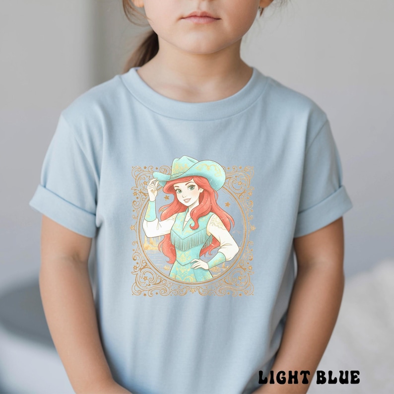 K&ouml;nnte beinhalten: Hellblaues T-Shirt mit einer Grafik eines rothaarigen Charakters mit Cowboyhut und t&uuml;rkisfarbenem Outfit, umrahmt von einem goldenen, verzierten Rand. Der Text "LIGHT BLUE" ist unten auf dem Shirt aufgedruckt.
