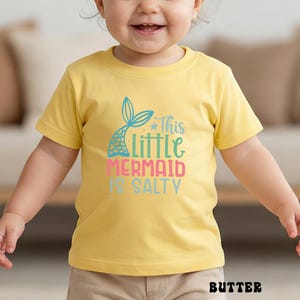Puede incluir: Una camiseta amarilla con el texto "This Little Mermaid Is Salty" en verde azulado, rosa y azul. La camiseta presenta un gr&aacute;fico de cola de sirena. La palabra "BUTTER" est&aacute; impresa en la parte inferior de la camiseta.