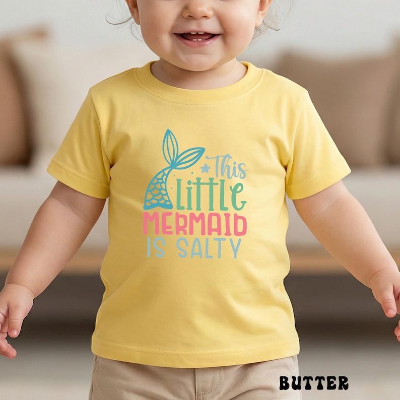 Puede incluir: Una camiseta amarilla con el texto "This Little Mermaid Is Salty" en verde azulado, rosa y azul. La camiseta presenta un gr&aacute;fico de cola de sirena. La palabra "BUTTER" est&aacute; impresa en la parte inferior de la camiseta.