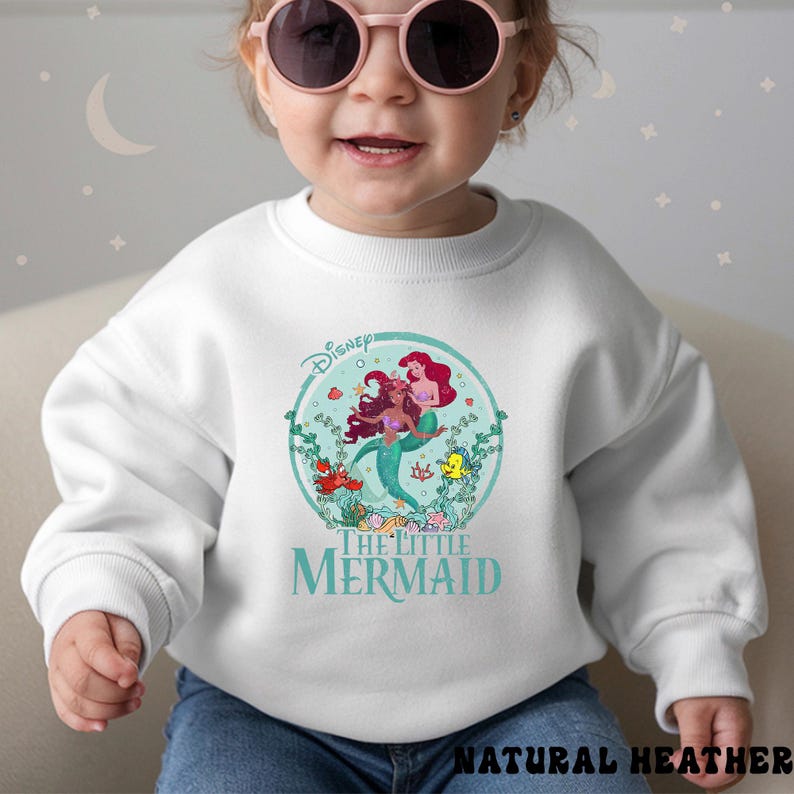 May include: White sweatshirt featuring a colorful graphic of Ariel and other characters from Disney's The Little Mermaid. The design is set within a circular frame with the Disney logo at the top. The words "The Little Mermaid" are printed below the graphic.