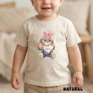 Peut inclure: T-shirt beige pour enfant avec un lapin de dessin anim&eacute; tenant un c&oelig;ur avec le mot "love". Le lapin porte un n&oelig;ud rose et une salopette bleue. Le mot "NATURAL" est imprim&eacute; en bas.