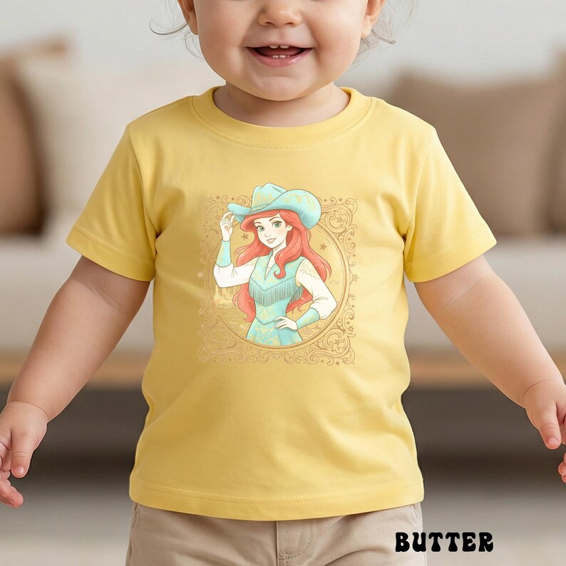 K&ouml;nnte beinhalten: Gelbes T-Shirt mit einer Cartoon-Illustration einer Frau mit Cowboyhut und t&uuml;rkisfarbenem Kleid, umrahmt von einem goldenen Oval. Das Wort "BUTTER" ist unten in Schwarz aufgedruckt. Geeignet f&uuml;r Kinder.