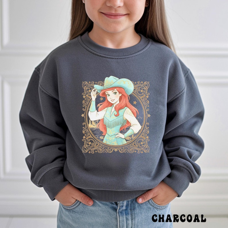 K&ouml;nnte beinhalten: Anthrazitgraues Sweatshirt mit einer Grafik einer Cowgirl mit roten Haaren und einem t&uuml;rkisfarbenen Hut. Die Cowgirl befindet sich in einem goldfarbenen, verzierten Rahmen. Das Wort "CHARCOAL" ist unten auf dem Sweatshirt wei&szlig; gedruckt.