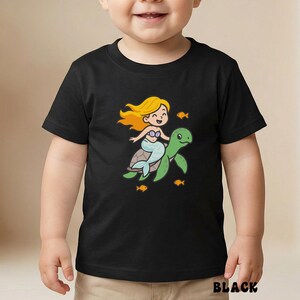 Puede incluir: Camiseta negra con una ilustraci&oacute;n de dibujos animados de una sirena montada en una tortuga verde, rodeada de peces naranjas. La sirena tiene el pelo rubio y una cola azul. La palabra "BLACK" est&aacute; impresa en la parte inferior.