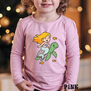 Puede incluir: Una camiseta rosa de manga larga con una sirena de dibujos animados montada en una tortuga verde, con peque&ntilde;os peces naranjas. La palabra "PINK" est&aacute; impresa en la esquina inferior derecha. La camiseta es para ni&ntilde;os.