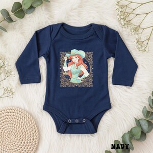 K&ouml;nnte beinhalten: Marineblauer Baby-Body mit langen &Auml;rmeln. Auf der Vorderseite befindet sich eine gerahmte Illustration einer rothaarigen Figur mit Cowboyhut und Fransenweste. Das Wort "NAVY" ist unten rechts im Bild aufgedruckt.