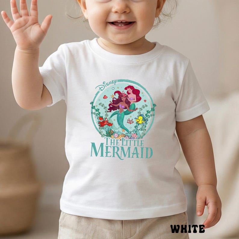 May include: White short-sleeved t-shirt featuring a colorful graphic of Ariel and another mermaid from Disney's The Little Mermaid. The design is set within a circular frame with aquatic elements. The text "THE LITTLE MERMAID" is printed below the graphic.