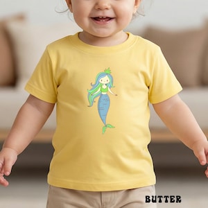 Puede incluir: Una camiseta amarilla de manga corta con una ilustraci&oacute;n de sirena de dibujos animados. La sirena tiene pelo azul y verde, un top verde y una cola azul. La palabra "BUTTER" est&aacute; impresa en negro en la parte inferior de la camiseta.