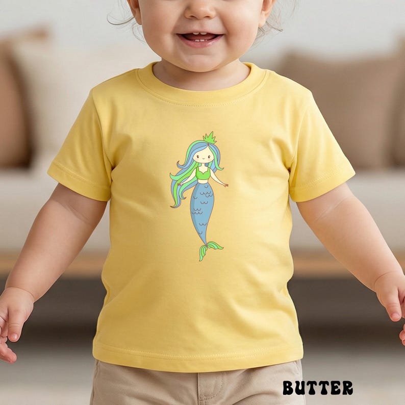 Puede incluir: Una camiseta amarilla de manga corta con una ilustraci&oacute;n de sirena de dibujos animados. La sirena tiene pelo azul y verde, un top verde y una cola azul. La palabra "BUTTER" est&aacute; impresa en negro en la parte inferior de la camiseta.