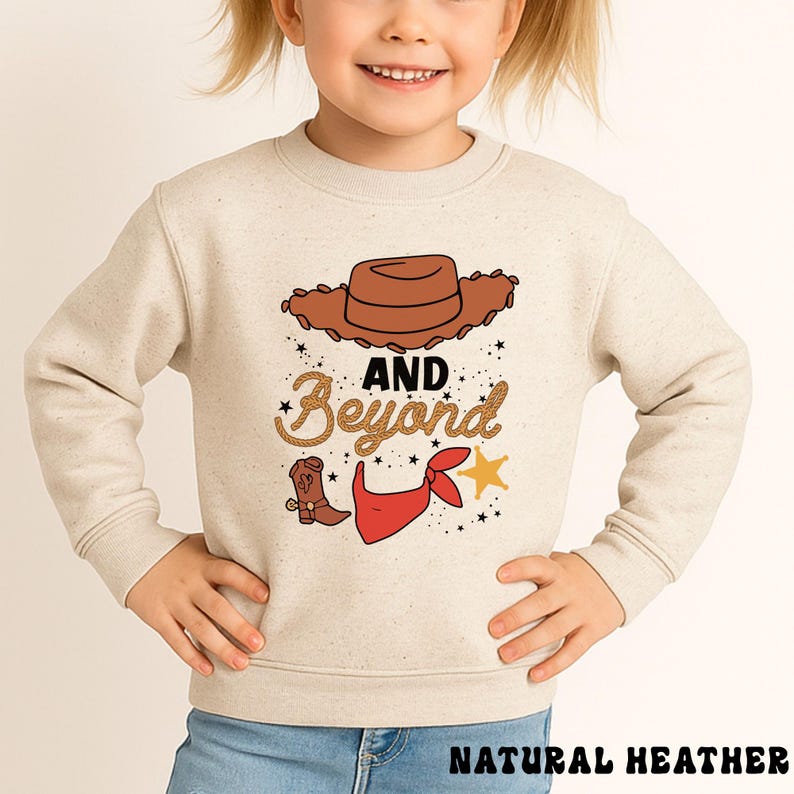 May include: A natural heather sweatshirt featuring a cowboy-themed graphic. The design includes a brown cowboy hat, the words "AND Beyond" in a decorative font, a cowboy boot, a red bandana, and a gold star. The text "NATURAL HEATHER" is at the bottom.