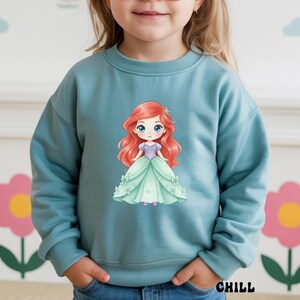 Puede incluir: Una sudadera de manga larga en color azul polvoriento con un gr&aacute;fico central de un personaje de dibujos animados con largo cabello rojo, corpi&ntilde;o morado y vestido de gala verde claro. El personaje tiene grandes ojos azules y una estrella.