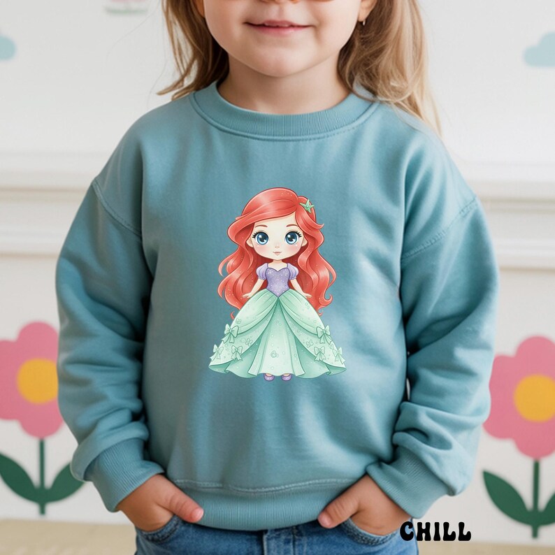Puede incluir: Una sudadera de manga larga en color azul polvoriento con un gr&aacute;fico central de un personaje de dibujos animados con largo cabello rojo, corpi&ntilde;o morado y vestido de gala verde claro. El personaje tiene grandes ojos azules y una estrella.