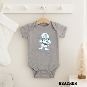 May include: A heather gray baby bodysuit featuring a cartoon illustration of Buzz Lightyear in a spacesuit. The design includes the text "Buzz Lightyear" in a teal color. The bodysuit is hanging on a wooden hanger.