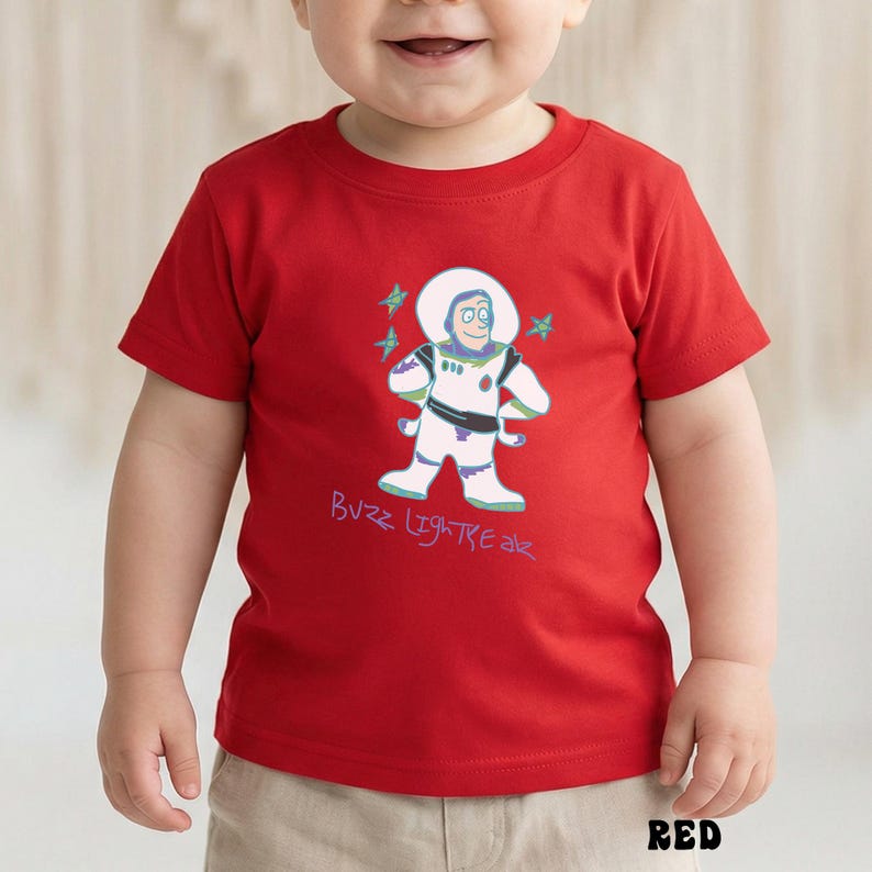 May include: A red t-shirt featuring a cartoon illustration of Buzz Lightyear with the text "Buzz Lightyear". The design includes a white spacesuit, green accents, and blue stars. The shirt is for babies and toddlers.