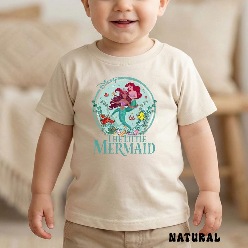 May include: A beige t-shirt featuring a colorful graphic of Ariel and other characters from Disney's The Little Mermaid. The design is within a circular frame, with the words "The Little Mermaid" below. The word "NATURAL" is printed at the bottom.