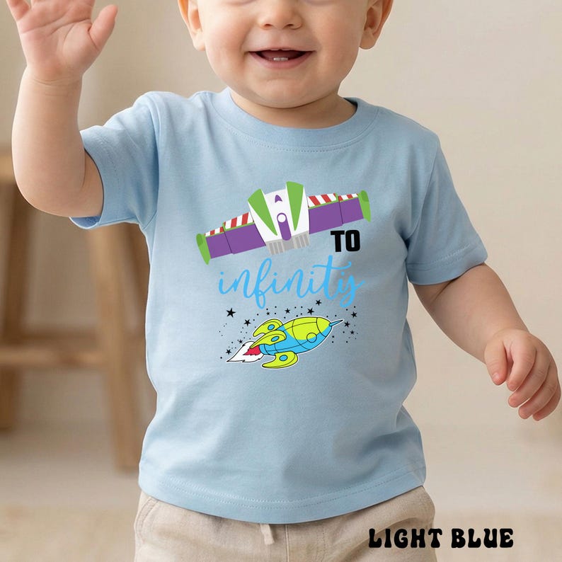 May include: Light blue toddler t-shirt with a colorful graphic of a spaceship and the phrase "To Infinity" in a playful font. The design includes a rocket and stars, perfect for a child's wardrobe.